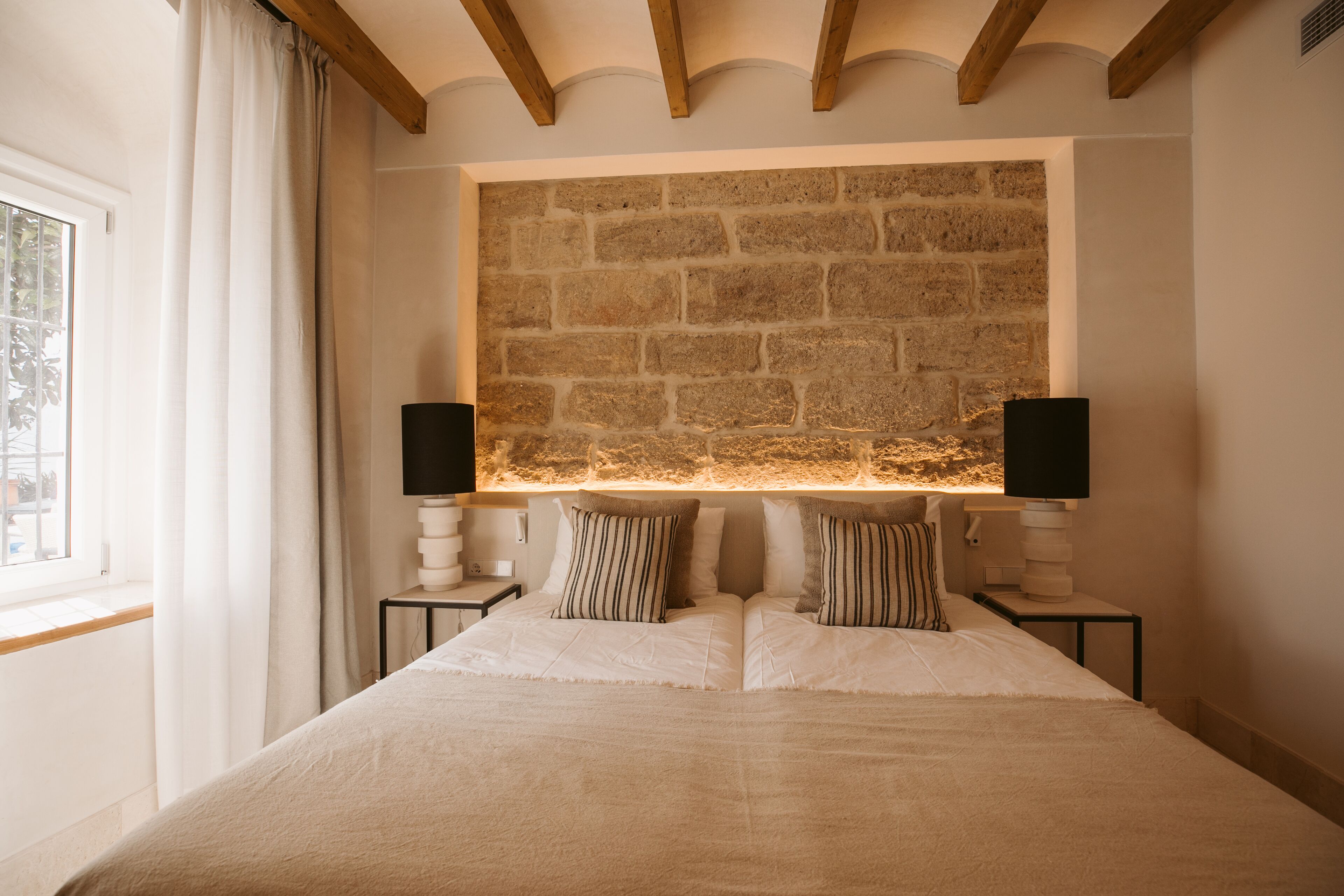 Ein Schlafzimmer mit Steinwand, einem großen Bett, zwei Nachttischen mit Lampen und einem Fenster mit Vorhängen.