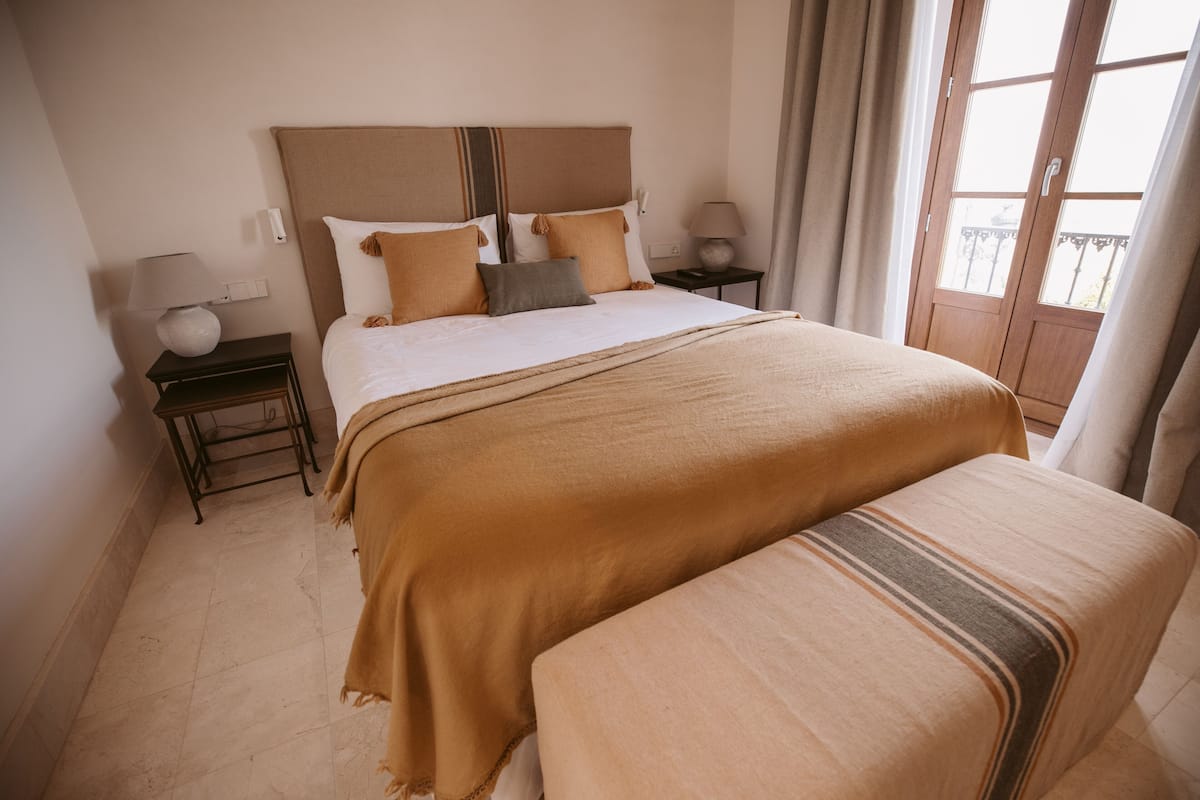 Habitación de hotel con una cama grande, dos mesitas de noche, un velador y una ventana con cortinas, con vista al balcón exterior.