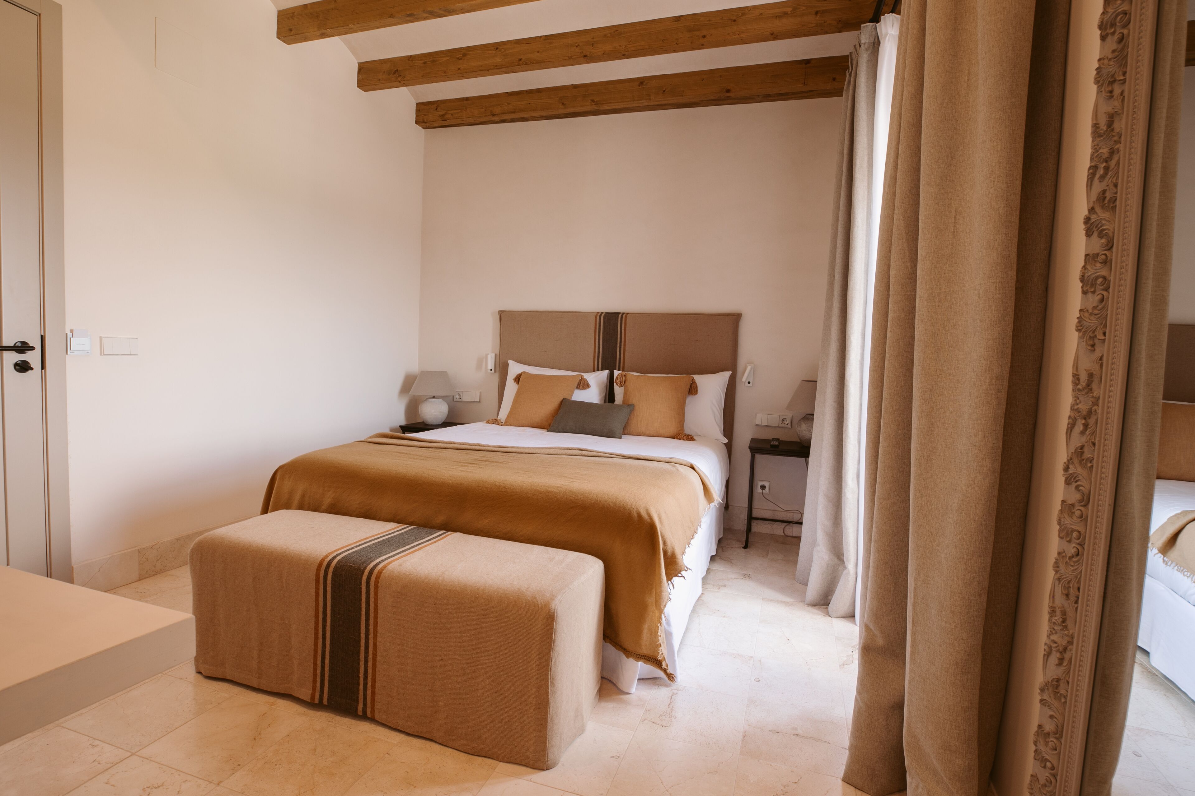 Un dormitorio con cama, mesitas de noche, un banco y cortinas.