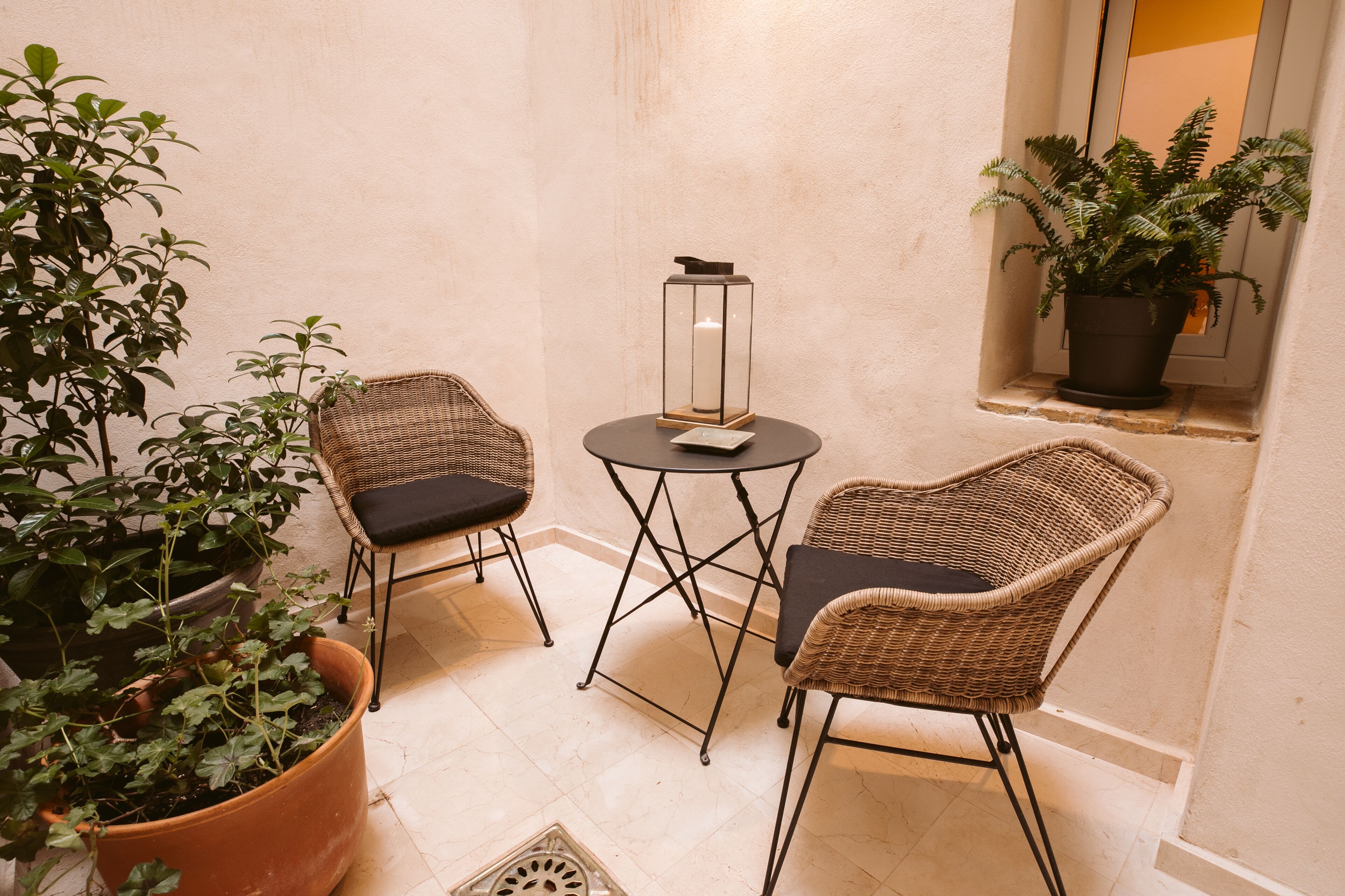 A small, cozy seating area with wicker chairs and a table, surrounded by potted plants.