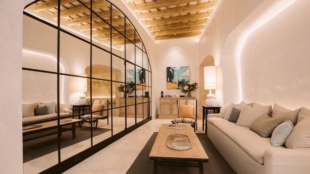 A modern living room with a curved glass partition, wooden ceiling beams, a sofa, a coffee table, and a kitchen area in the background. - Casa Aura (Vejer de la Frontera)