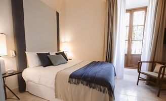 Una cama bien tendida con una manta azul, una lámpara de noche y una silla en una habitación luminosa con un ventanal y cortinas.