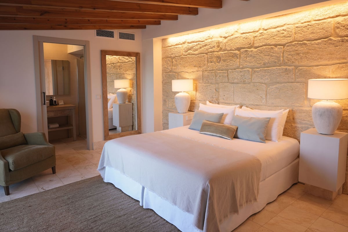 Un dormitorio con una cama grande, dos mesitas de noche con lámparas, un sillón verde y una pared de piedra.