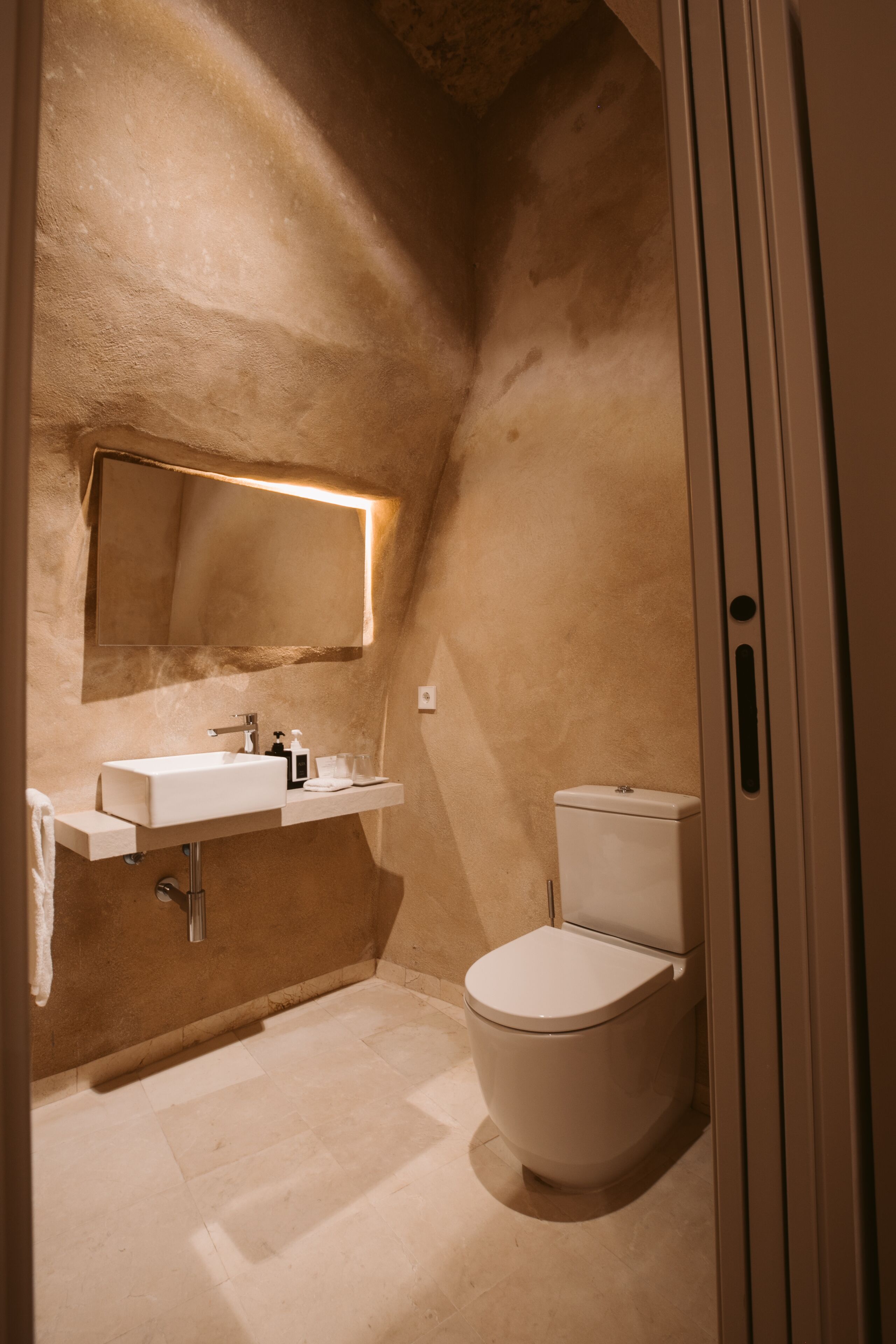 A compact bathroom with a white sink, mirror, and toilet, featuring a textured wall and a small shelf.