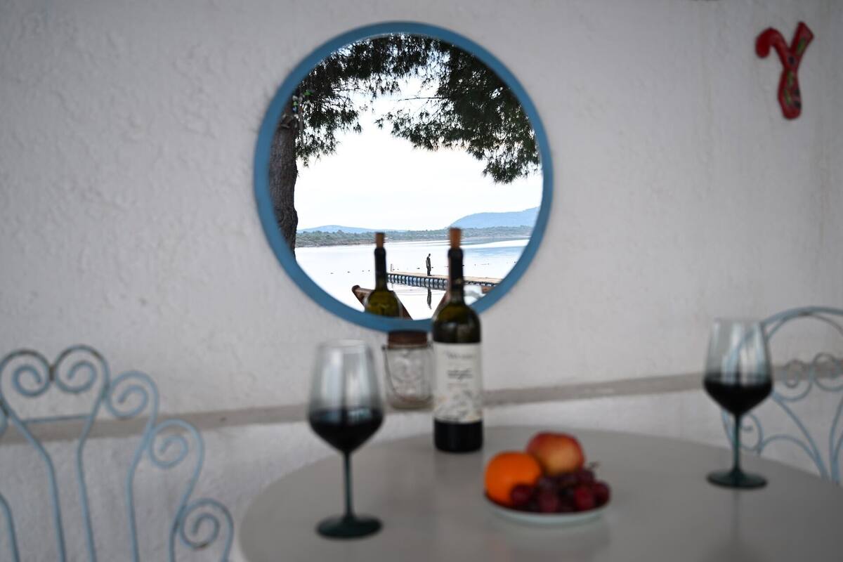 A round mirror reflecting a lake view with mountains, two wine bottles, and a plate of fruit on a table.