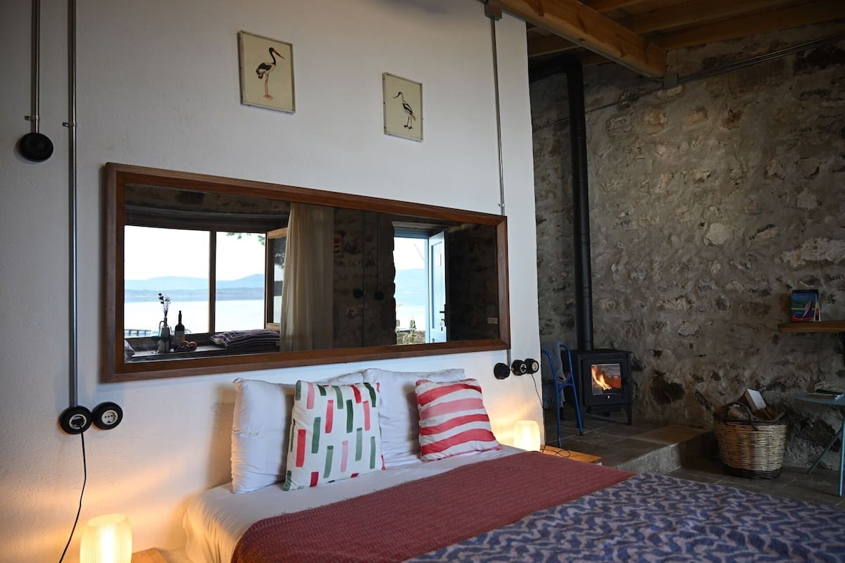 A bedroom with a large mirror, a bed with patterned pillows, a stone wall, and a wood-burning stove.