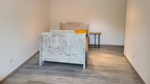 Um quarto com uma cama de madeira rústica, uma mesinha de cabeceira pequena e uma parede branca simples.