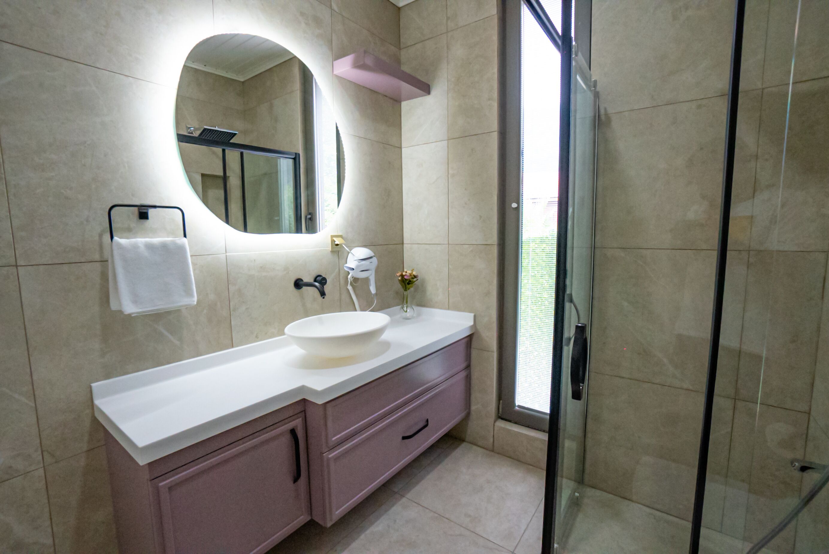 A modern bathroom with a large oval mirror, a white sink, a pink vanity, and a glass shower enclosure.