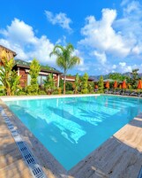 Una piscina con agua azul cristalina, rodeada de plantas tropicales y un edificio de madera con techo rojo.