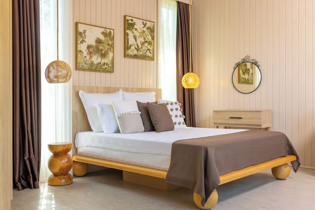 Uma cama com estrutura de madeira, roupa de cama branca e manta marrom. Há duas obras de arte penduradas na parede e um espelho redondo.