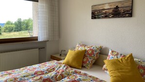 A bedroom with a bed, colorful floral bedding, a window with curtains, a bedside table, and a painting on the wall.