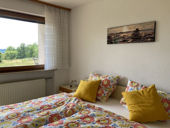 A bedroom with a bed, colorful floral bedding, a window with curtains, a bedside table, and a painting on the wall.
