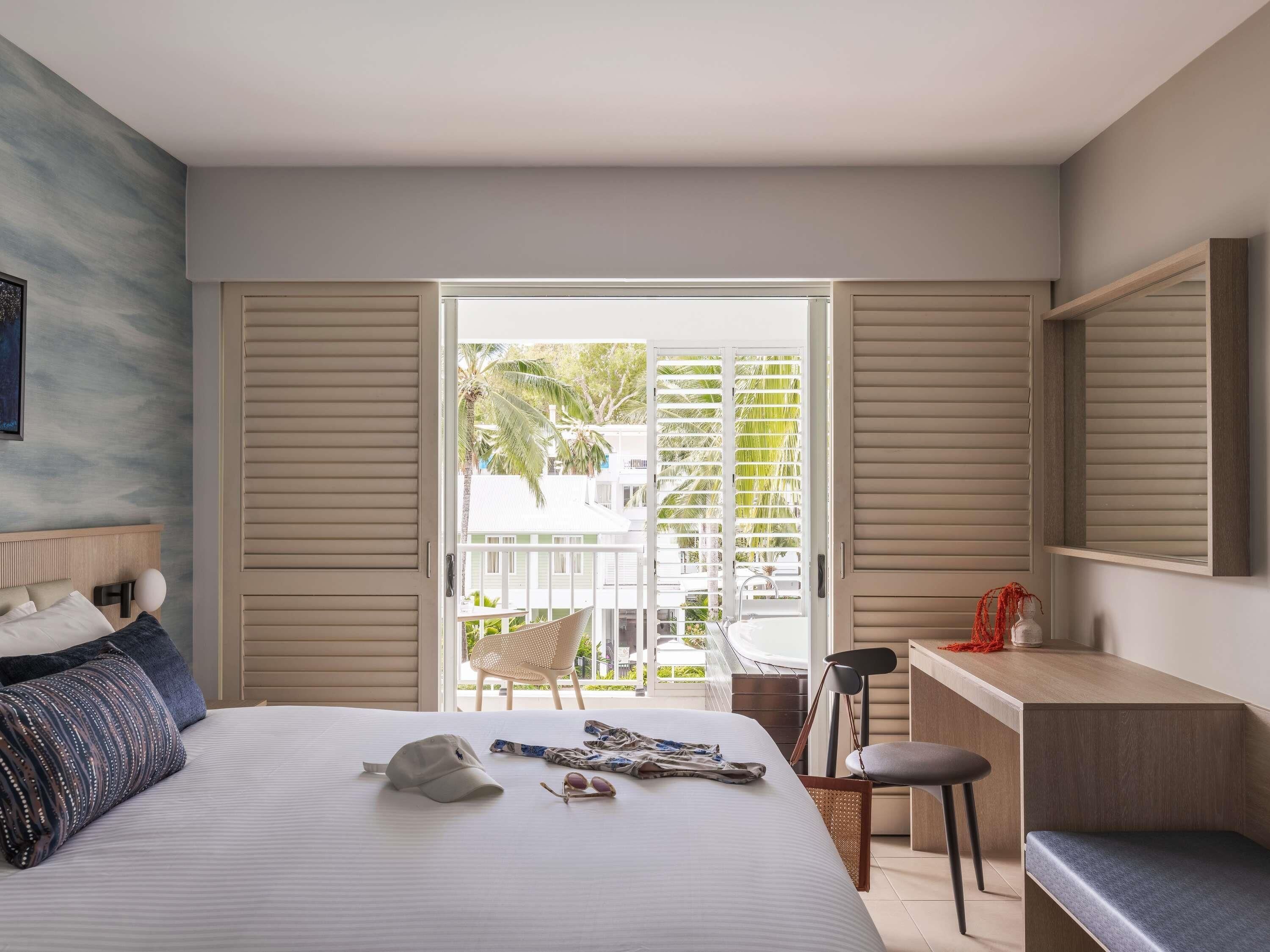 Habitación de hotel con cama, escritorio, silla y balcón con vistas a palmeras y edificios.