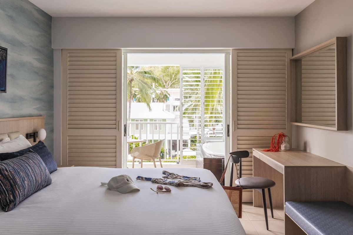 Habitación de hotel con cama, escritorio, silla y balcón con vistas a palmeras y edificios.