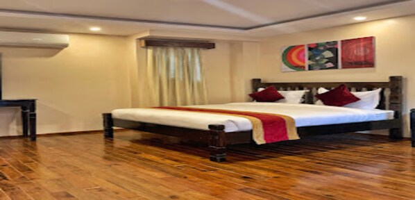 A hotel room with a wooden floor, a large bed with white bedding and red pillows, a window with curtains, and a wall with a colorful artwork.