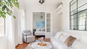 Un salon moderne avec un canapé blanc, une table basse ronde sur laquelle repose un plateau, un fauteuil gris et un lit à l’arrière-plan.