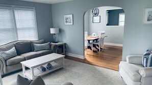 A living room with a sofa, coffee table, and chairs, a dining area with a table and chairs, and a hallway leading to another room.