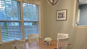 A dining area with a wooden table and white chairs, a chandelier, and a window with blinds.