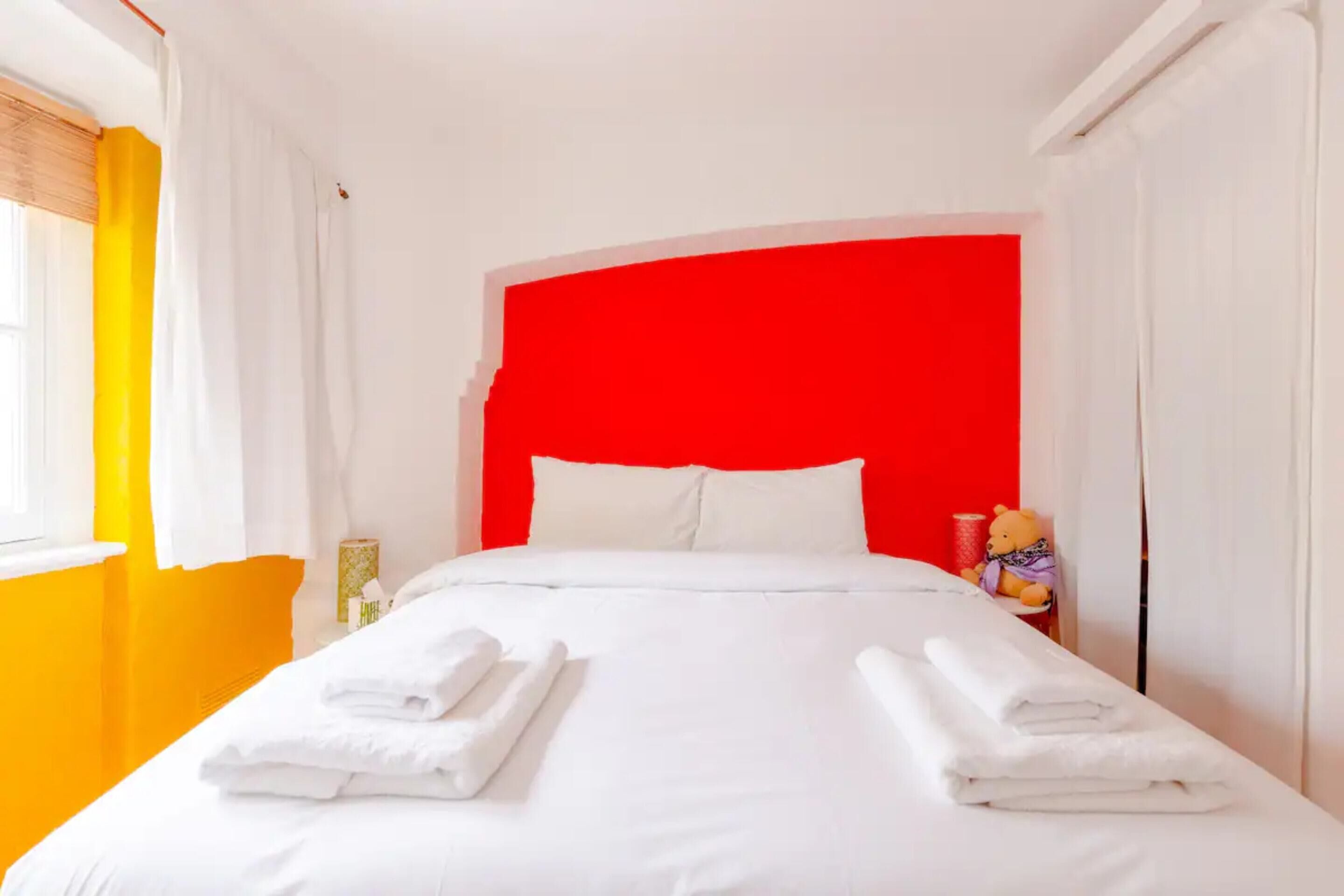 Habitación de hotel con una pared de acento roja, una cama bien hecha con ropa blanca y un osito de peluche en la mesita de noche.