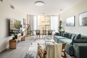 A modern living room with a sectional sofa, a dining table with chairs, a TV, and a balcony with a view of a building. - Spacious & Bright 3BR Apt with City View (Tel Aviv)