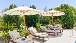 Zona de piscina con sillones de descanso y sombrillas, rodeada de abundante vegetación.