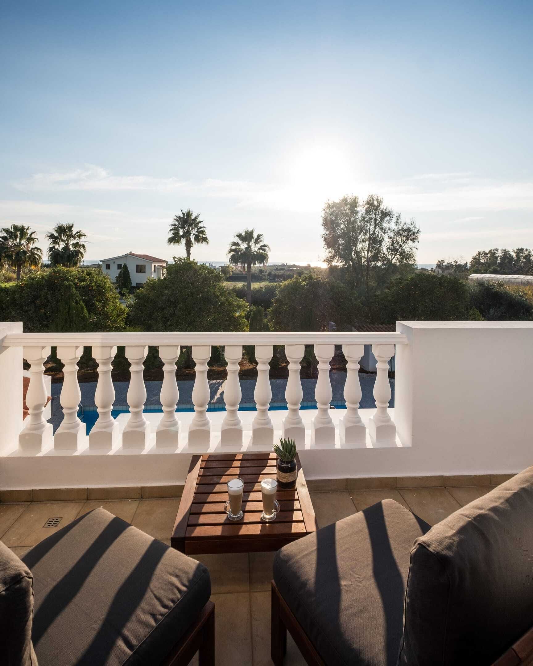 Un balcón con barandilla blanca, dos sillones con cojines, una mesita con dos vasos y vistas al mar y a la vegetación.