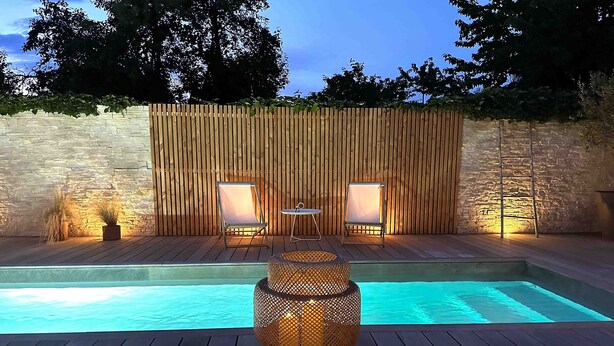 A pool area with a wooden deck, two lounge chairs, a small table, and a decorative lamp. - Maison de Charme (Valence)