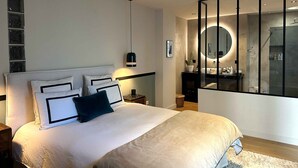 A modern bedroom with a large bed, bedside tables, a nightstand, a mirror, and a view into the bathroom. - Maison de Charme (Valence)
