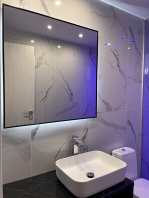 A modern bathroom with a large mirror, marble tiles, a white sink, and a toilet. - Double Bee Villa with private pool (Oropedio Lasithiou)
