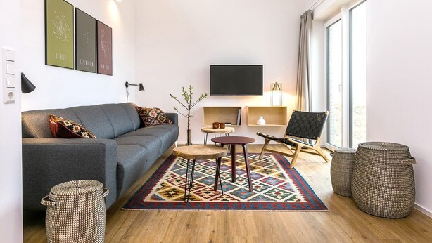 Ein kompakter Wohnraum mit grauem Sofa, einem kleinen runden Tisch, einem schwarzen Stuhl und einem farbenfrohen Teppich.