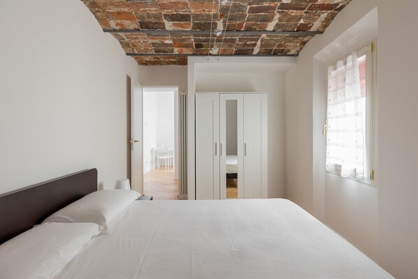 A bedroom with a brick ceiling, white walls, a bed with white linens, a wardrobe, and a window with sheer curtains.