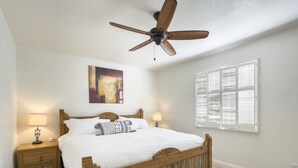 A bedroom with a wooden bed, two bedside tables with lamps, a ceiling fan, a window with shutters, and a painting on the wall. - Gorgeous and private ranch on expansive 1.2 acres (Scottsdale)