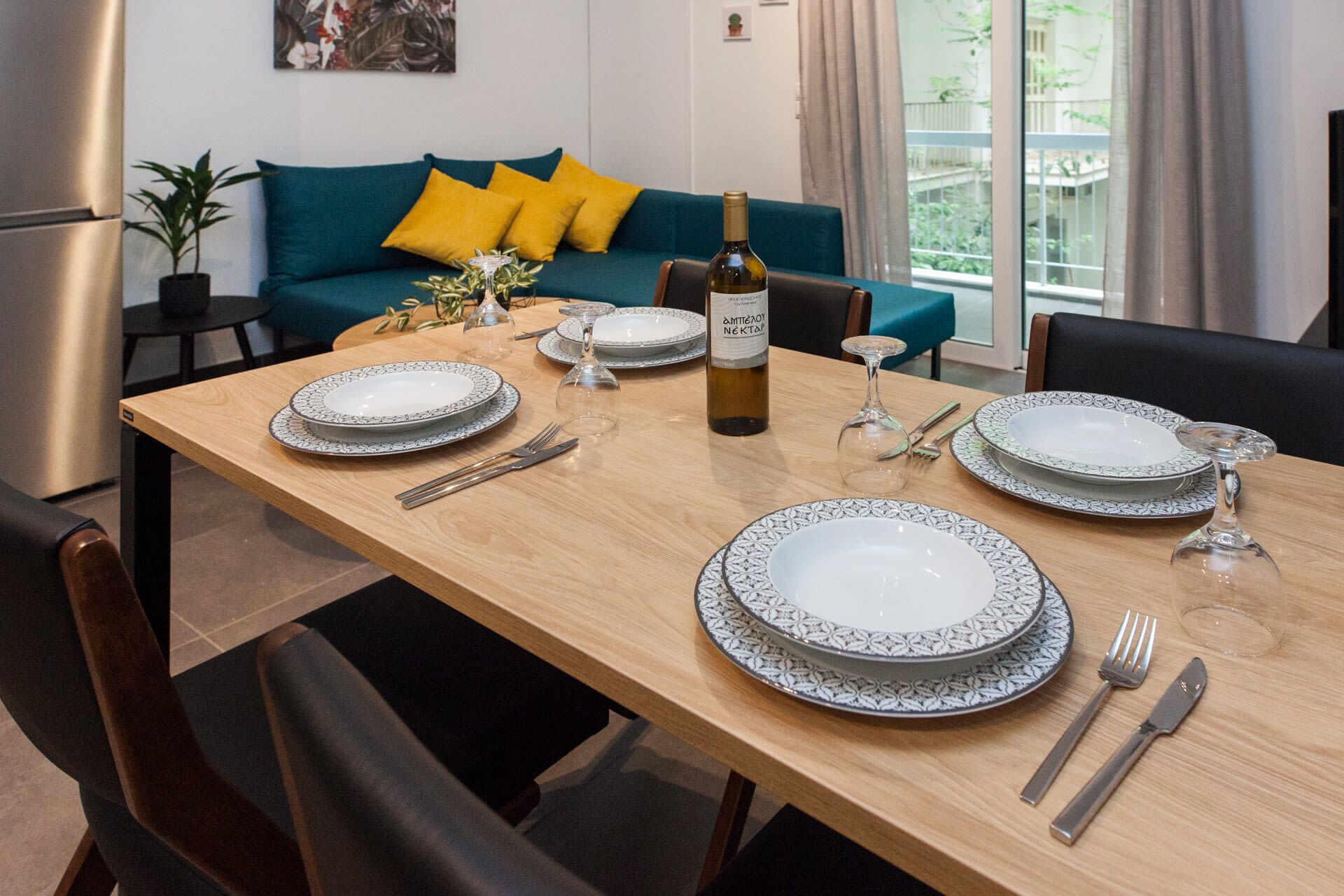 Une table à manger dressée avec des assiettes, des couverts et une bouteille de vin, avec un canapé et une plante en arrière-plan.