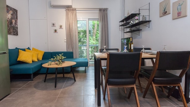 A modern living room with a blue sofa, yellow cushions, a coffee table, and a dining area with a wooden table and chairs. - Elegant Urban Escape Apt in Kolonaki (Athens)
