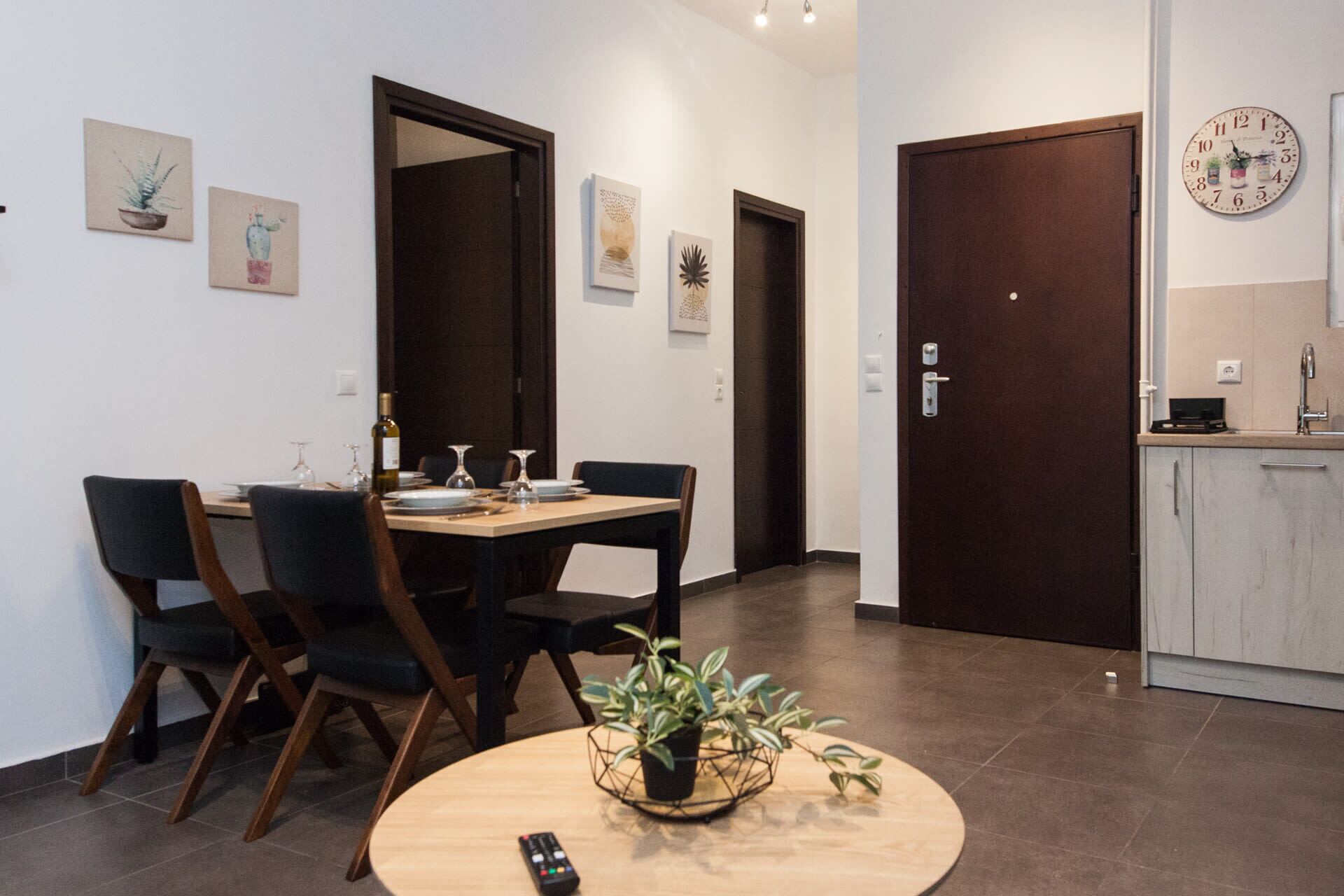 Un espace repas moderne avec une table en bois dressée pour quatre personnes, entourée de chaises. On aperçoit une petite table ronde avec une plante au premier plan. La pièce comprend un coin cuisine avec un évier et des placards, ainsi qu’une horloge au mur.