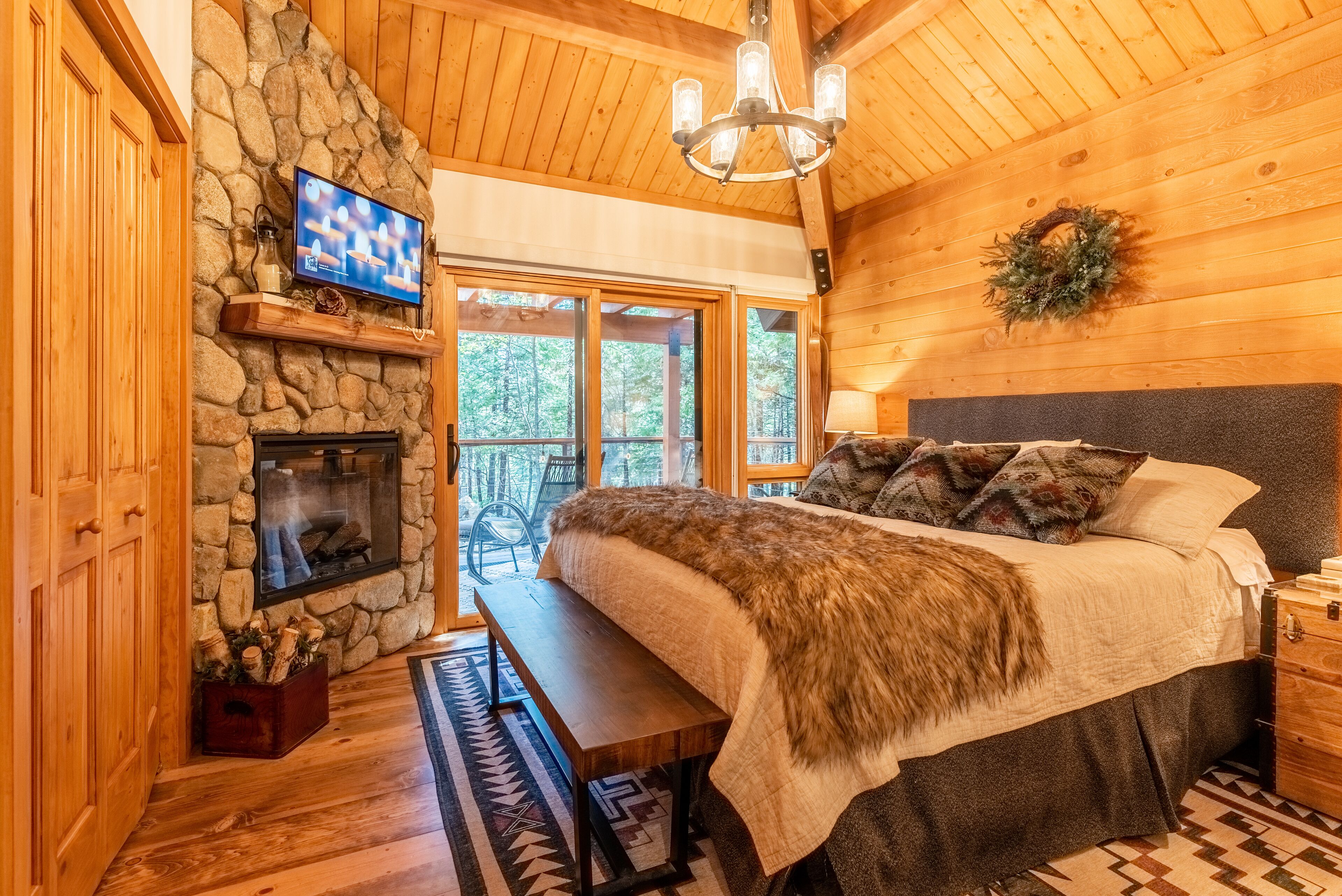 A cozy log cabin bedroom with a stone fireplace, a large bed with a fur throw, and a television mounted on the wall.