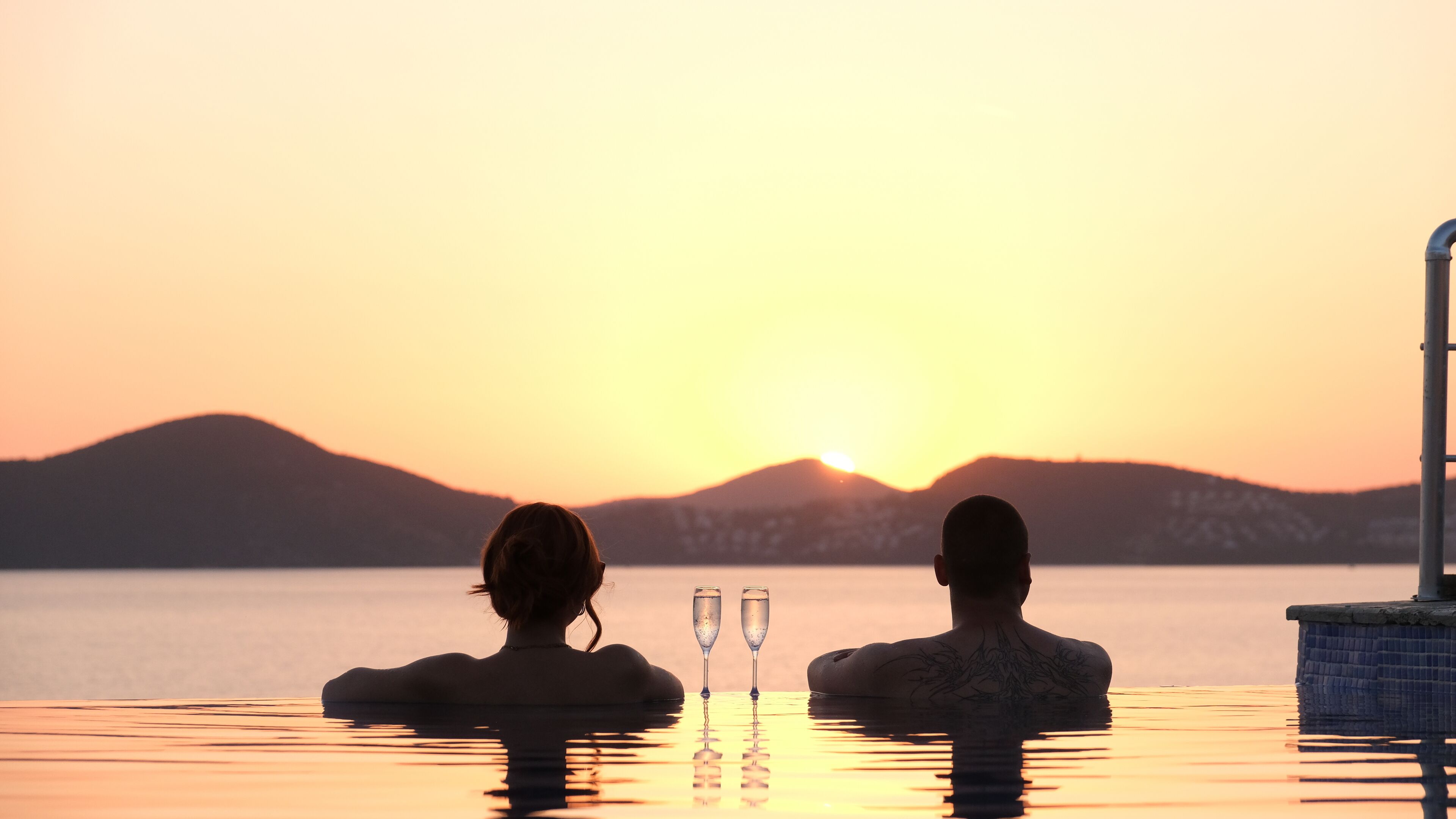 Two people in a swimming pool at sunset with mountains in the background.