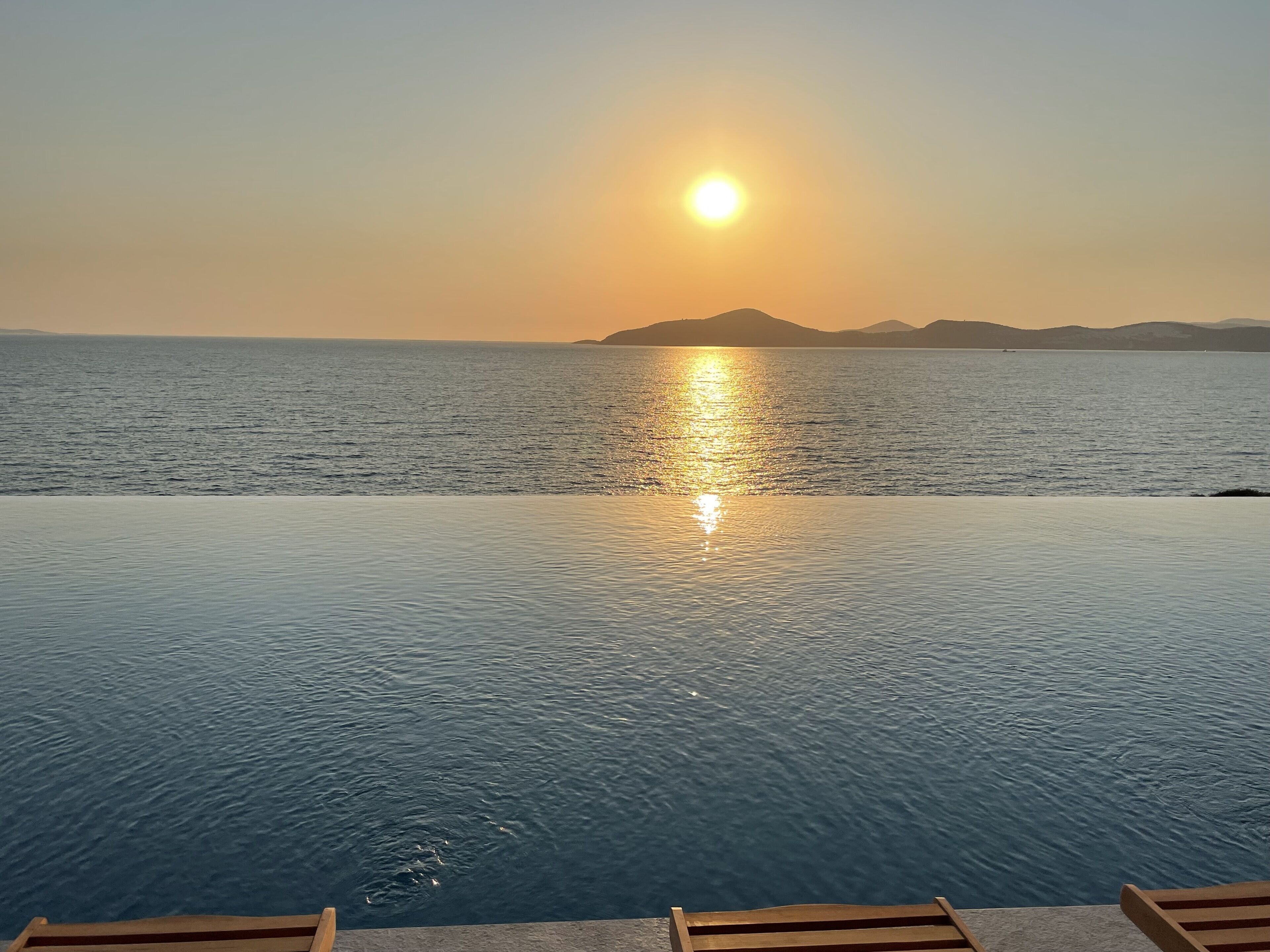 A poolside view with a sunset over the water, wooden deck chairs, and distant mountains.