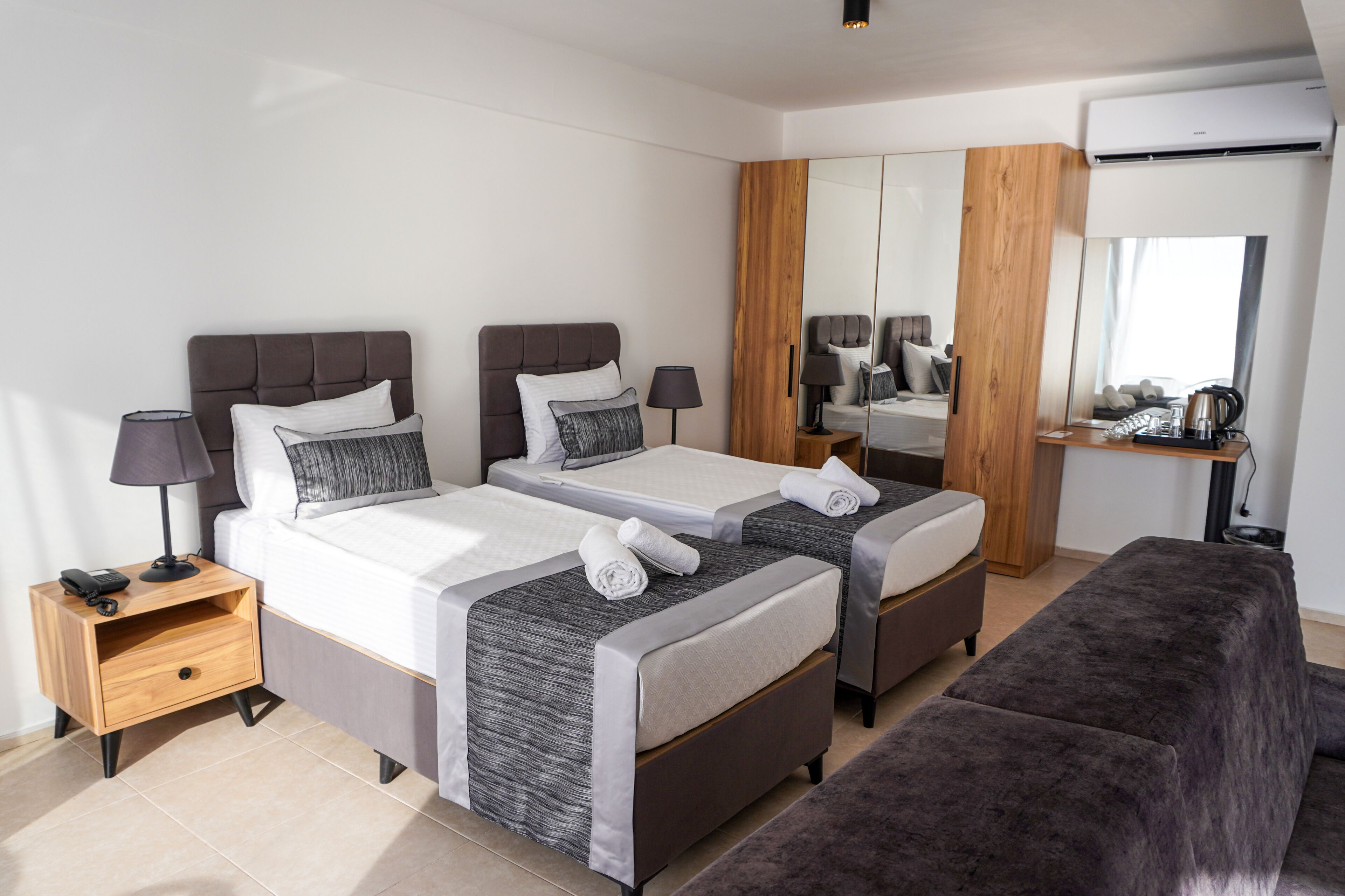 A modern hotel room with two beds, a wooden wardrobe, a mirror, and a small seating area.