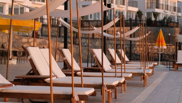 A row of wooden lounge chairs with white cushions, arranged on a tiled patio with a stone wall in the background.