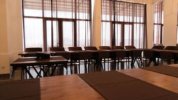 A conference room with multiple tables and chairs, large windows with blinds, and a wooden floor.