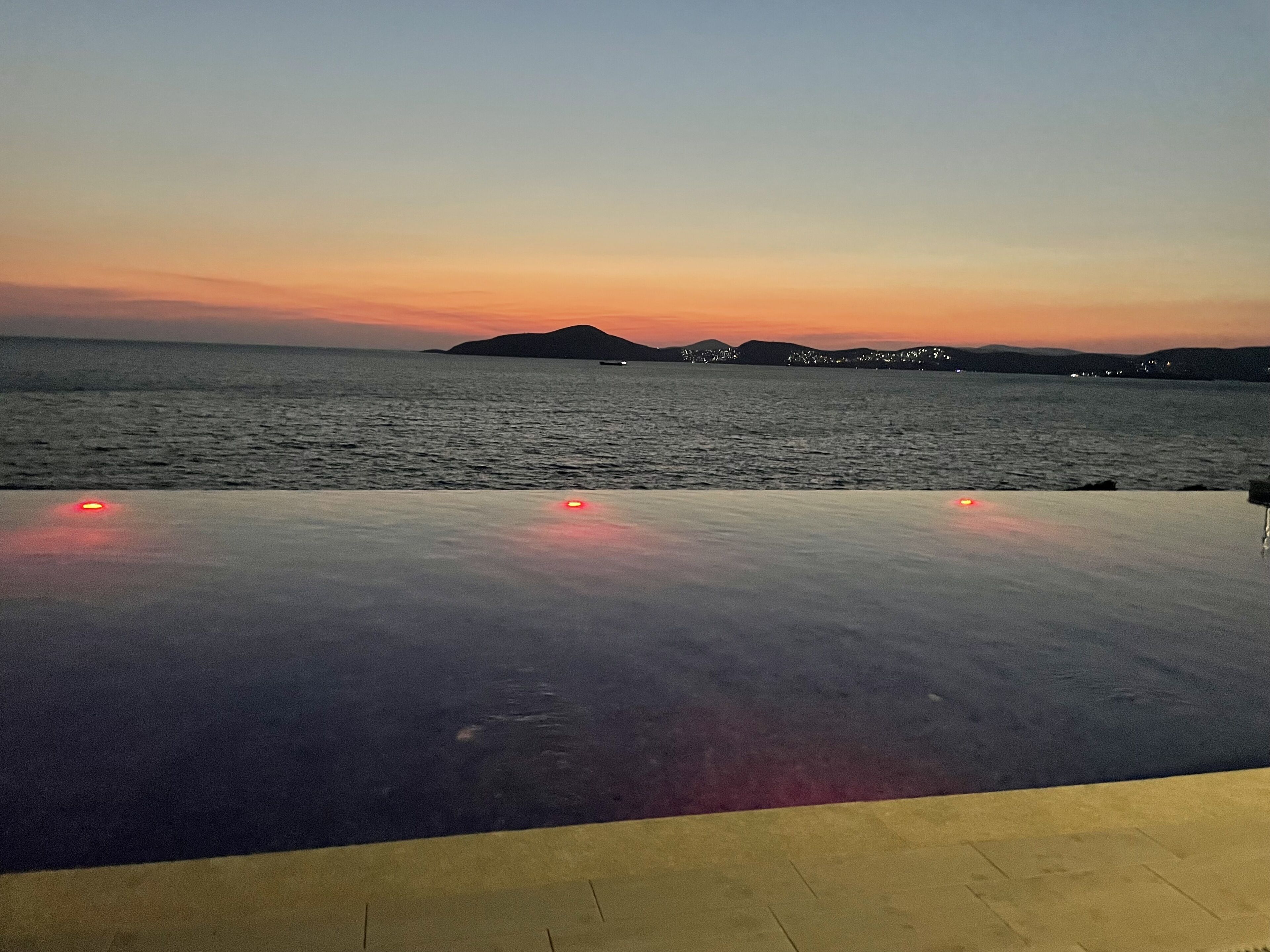 An outdoor pool area with a view of the sea and distant hills during sunset.