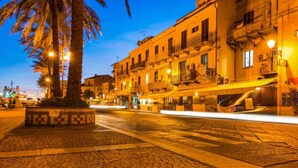 A cobblestone street at dusk with illuminated buildings, a palm tree, and a tiled planter. - Affittimoderni La Maddalena - MADA28 (La Maddalena)