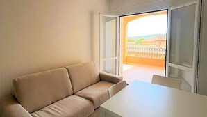 A living room with a beige sofa, a white table, and chairs. There is a sliding glass door leading to an outdoor balcony with a view of a building and a fence. - Affittimoderni La Maddalena - MADA28 (La Maddalena)