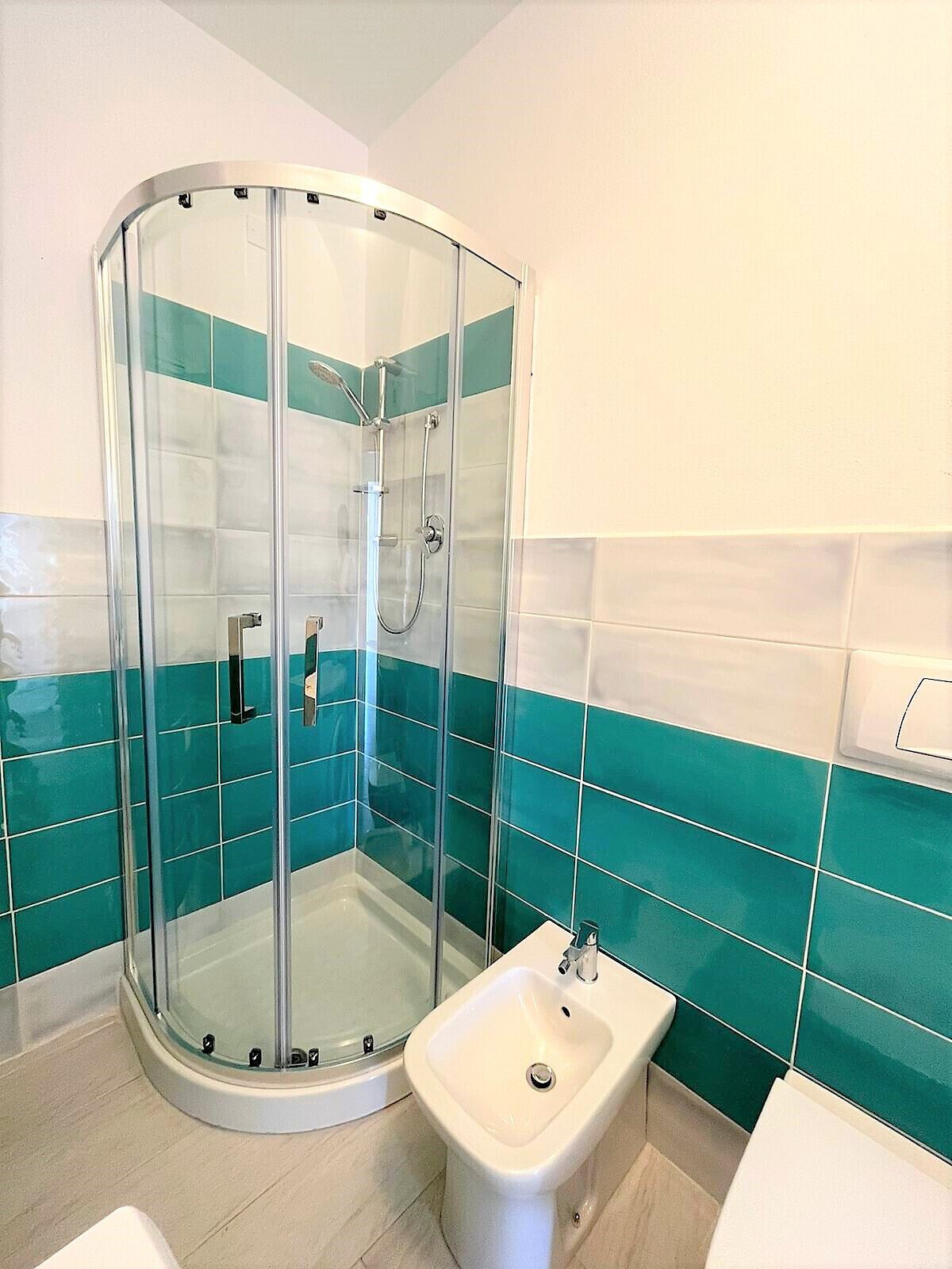 A modern bathroom with a glass-enclosed shower, a bidet, and teal and white tiled walls.