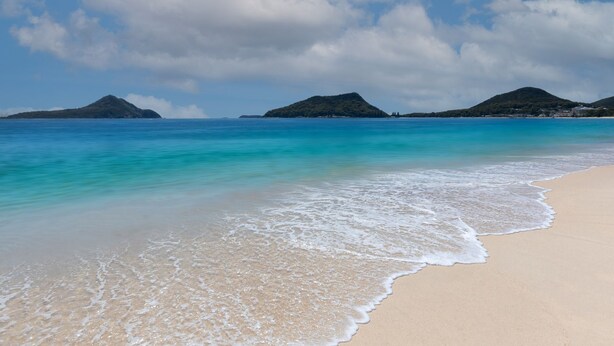 A beach with clear turquoise water, gentle waves, and distant mountains.
