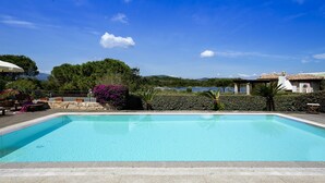 A rectangular swimming pool with a wooden deck in the foreground, surrounded by lush greenery and a clear blue sky.