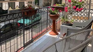 Un balcón con una silla de mimbre, plantas en macetas y vistas a una calle con coches estacionados.