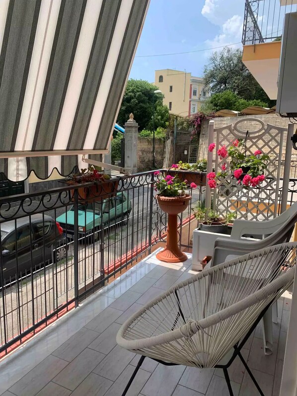 A balcony with a wicker chair, potted plants, and a view of a street with parked cars.