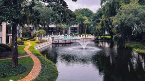 A serene courtyard with a fountain, a pool, and a pathway leading to a building with balconies. - Home away from Home. Fresh, Bright Condo just steps away from the beach (Hilton Head Island)